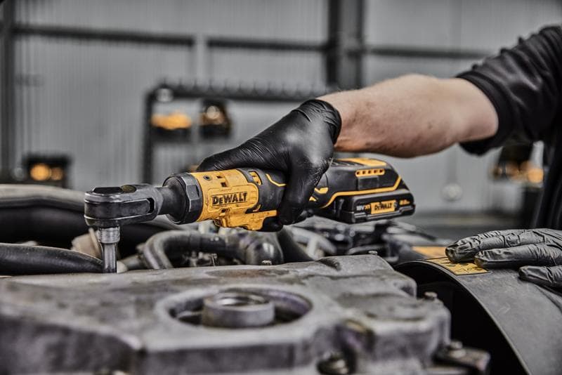 A person wearing black gloves uses a DEWALT cordless ratchet wrench to work on an engine component in an industrial setting.