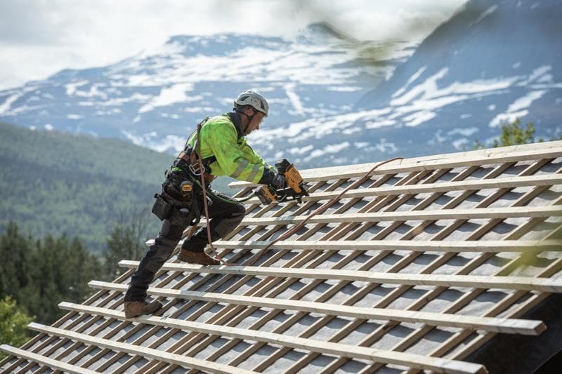 Worker using a DEWALT nailer on a sloped roof outdoors, wearing safety gear, with mountains in the background.
