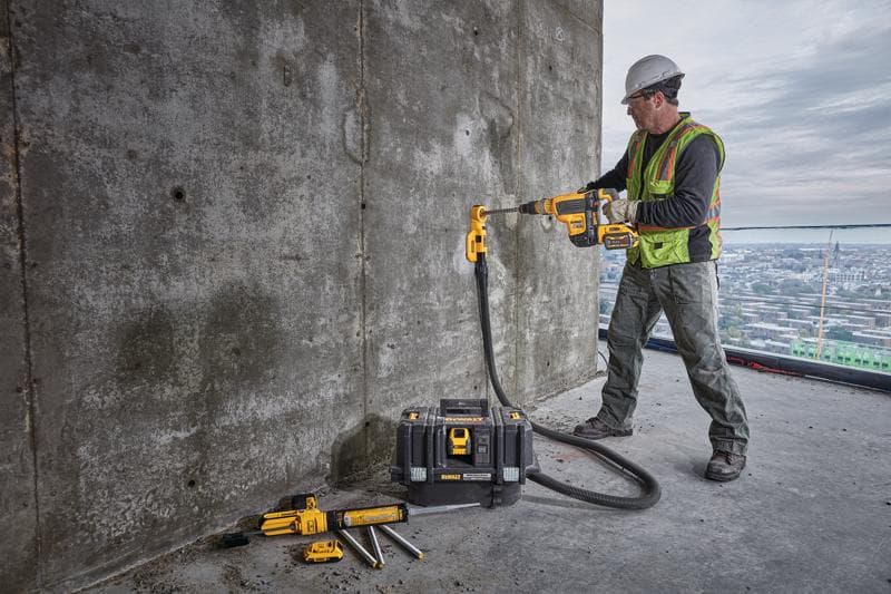 A construction worker wearing a safety vest and helmet is using a DEWALT cordless drill with dust extraction attachment to drill into a concrete wall. The worker is standing on a large building with a cityscape in the background. Several DEWALT tools and a tool case are placed on the ground nearby.