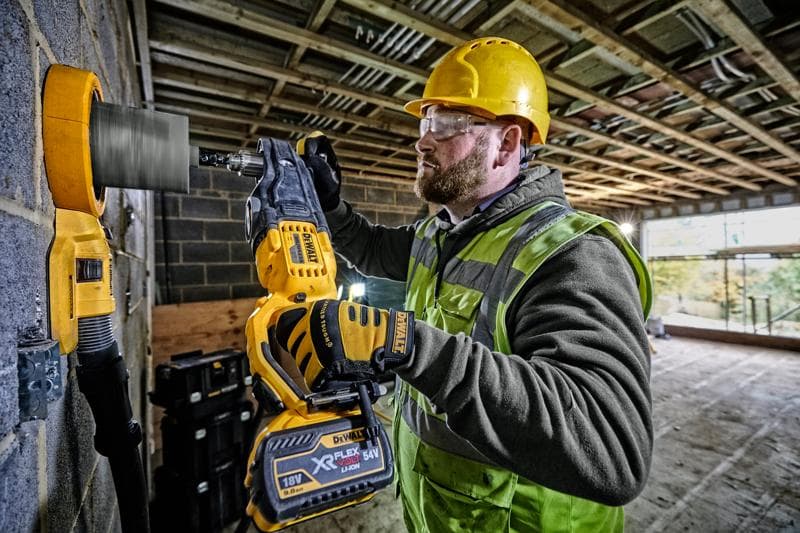 A construction worker wearing a yellow hard hat and high-visibility vest uses a DEWALT DCD470N cordless drill with a hole saw attachment to drill into a concrete wall at a building site.