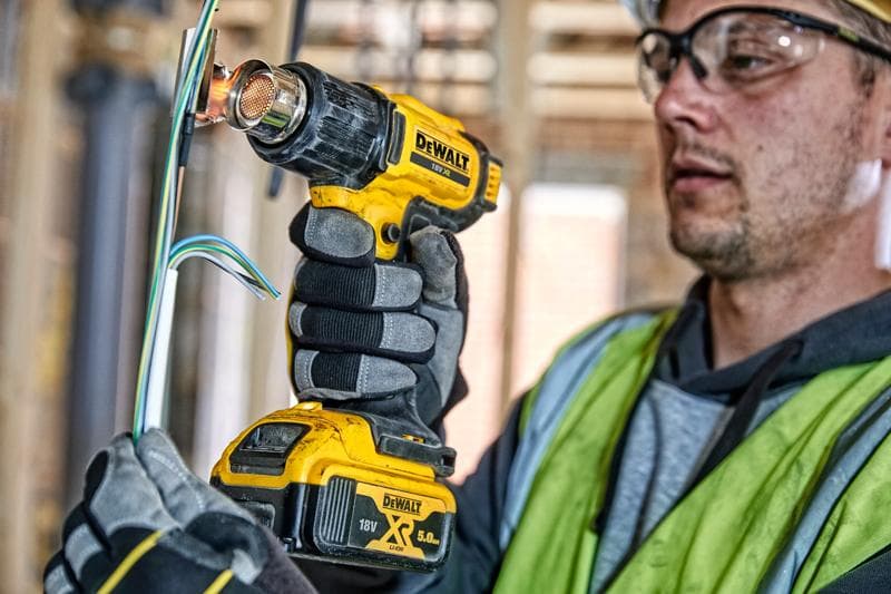 A person wearing work gloves and a safety vest uses a DEWALT DCE530N cordless heat gun to apply heat to electrical wires at a construction site. The person's face is blurred for privacy.