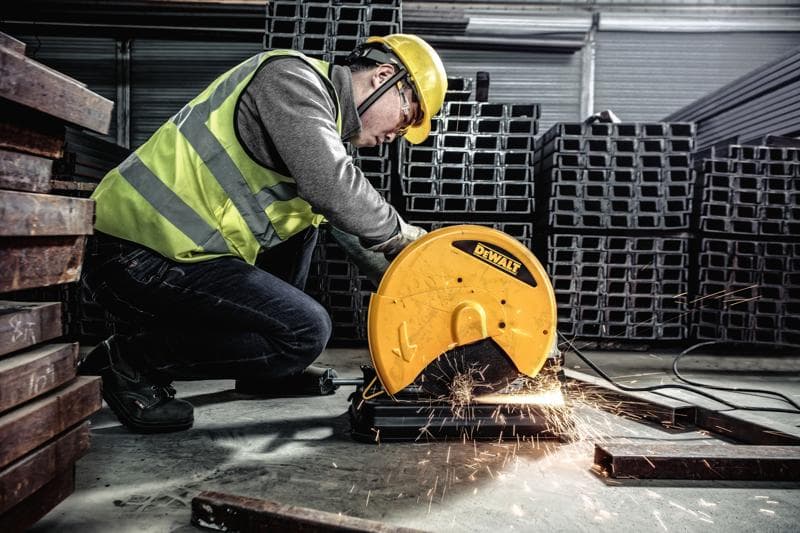 A person wearing safety gear operates a DEWALT metal cutting saw, model D28730, to cut metal bars in an industrial setting. Sparks fly from the cutting area.