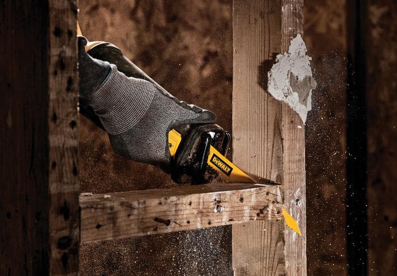 A close-up of a hand wearing a gray glove using a DEWALT reciprocating saw to cut through a wooden beam, with sawdust spraying in the air.