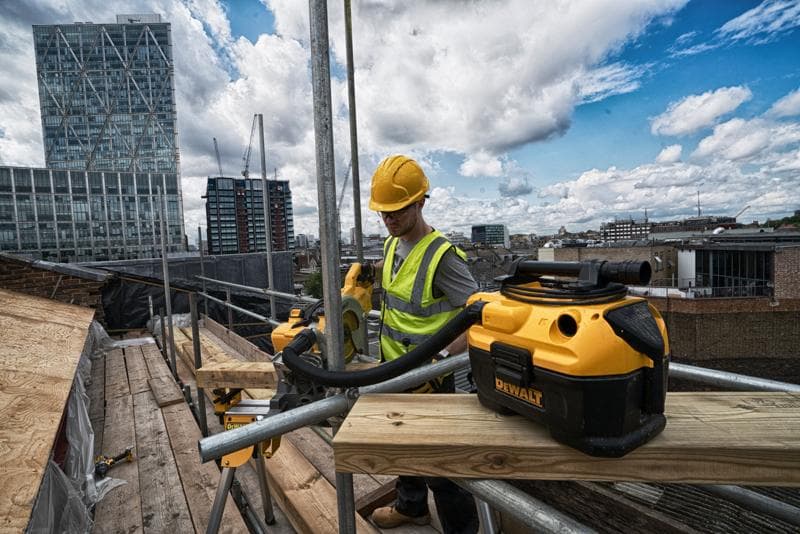 DEWALT DCV584 vacuum on a rooftop construction site with a worker in safety gear and city buildings in the background.