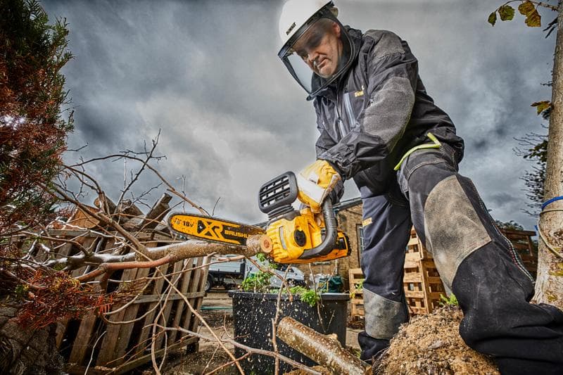 Person wearing safety gear uses a DEWALT chainsaw to cut branches outdoors on a cloudy day.