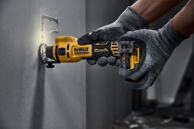 DEWALT cordless rotary tool cutting into drywall, operated by gloved hands.