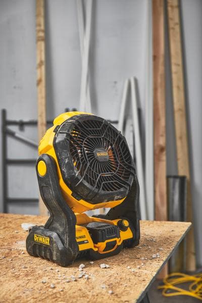 Přenosný akumulátorový ventilátor DEWALT (model DCE512B) je umístěn na dřevěné pracovní desce v dílně, na pozadí jsou stavební materiály.