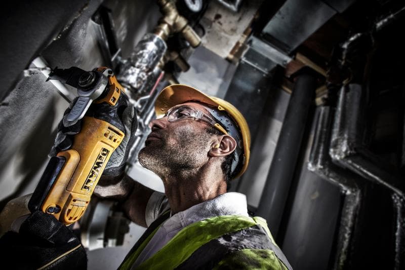 Worker uses a DEWALT DWE315 tool to cut wall pipe, wearing safety helmet and vest in industrial setting.