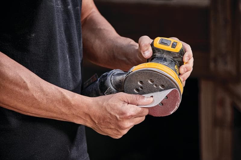 A person holding a DEWALT cordless random orbital sander, model DCW210B, attaching a sanding disc to the tool.