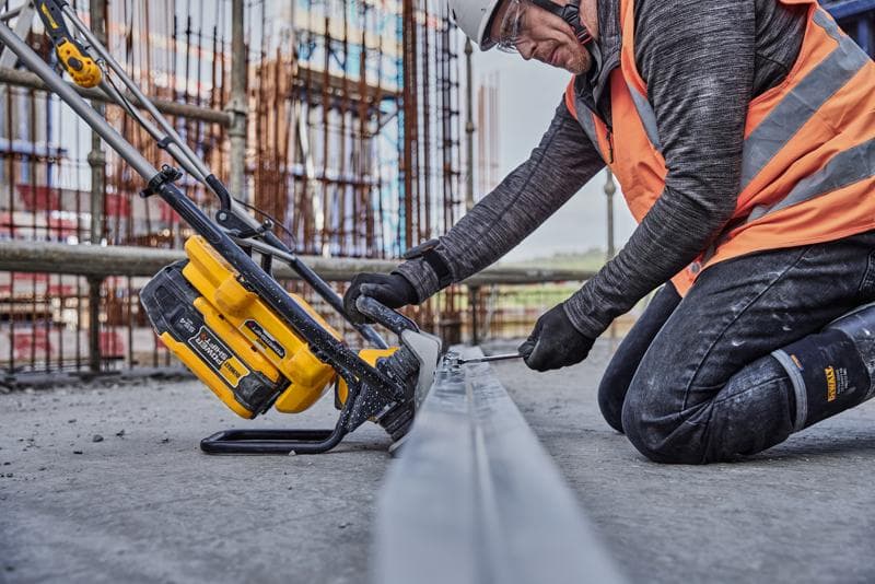 Worker uses yellow DEWALT tool to saw metal at construction site, wearing safety gear and orange vest.