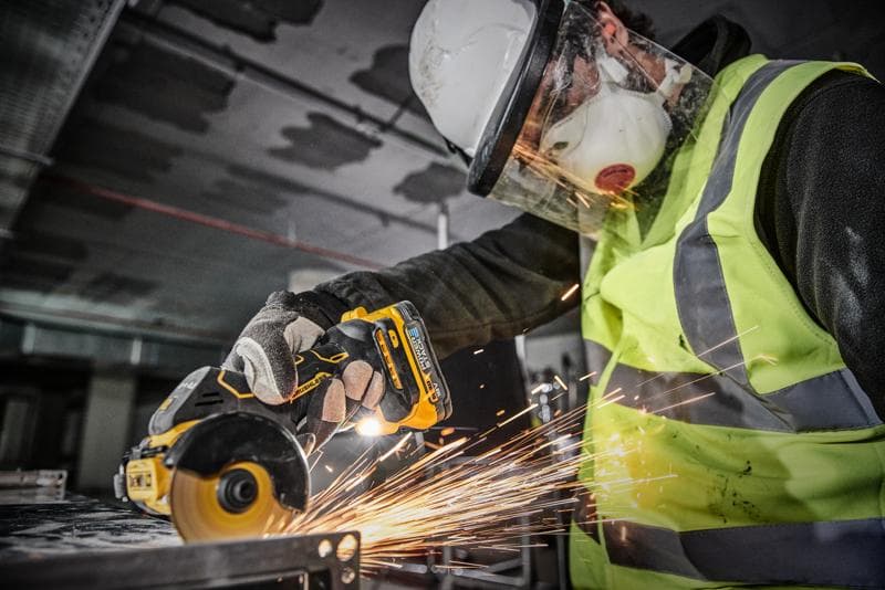 Osoba s bezpečnostním vybavením, včetně helmy, obličejového štítu, masky, rukavic a reflexní vesty, používá akumulátorový nástroj DEWALT DCS438N k řezání kovu, přičemž vznikají jiskry.