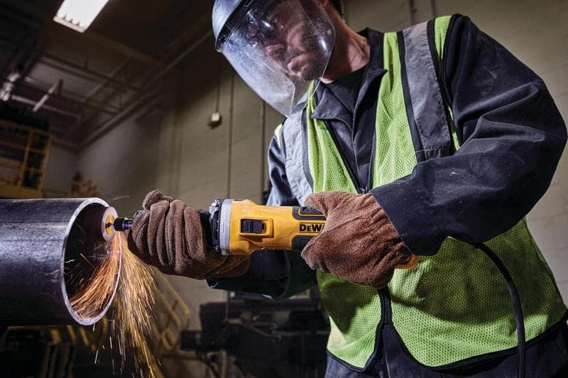 Worker using a DEWALT angle grinder to cut a metal pipe, sparks flying, wearing gloves and safety gear indoors.