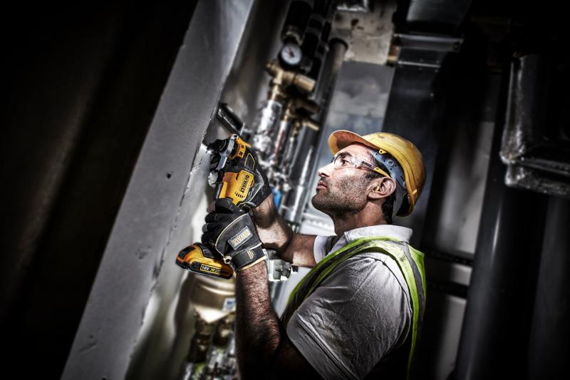 Worker uses a DEWALT DCS355D2 power tool on a wall at a construction site, wearing safety gear and gloves.