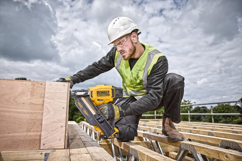 Person uses a DEWALT nail gun DCN930N on wooden boards at an outdoor construction site, wearing safety gear.