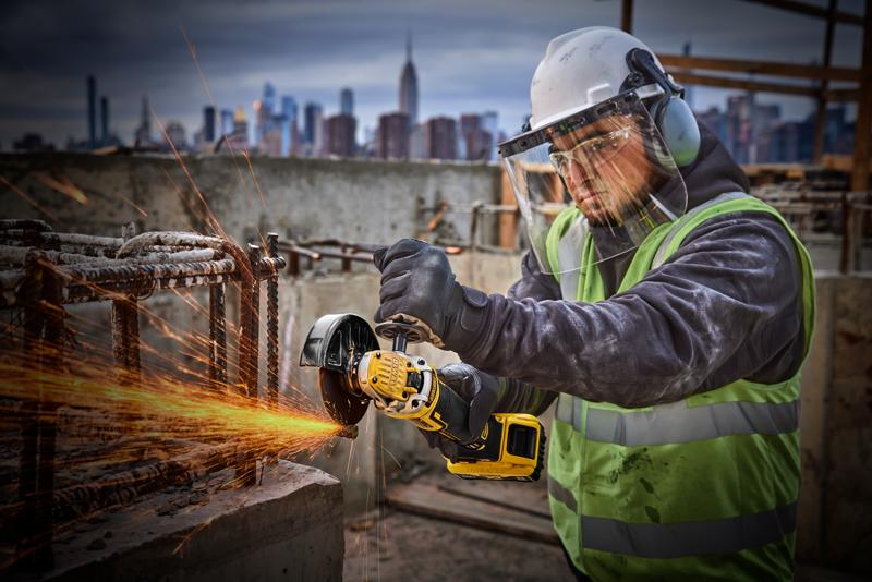 Worker uses DEWALT DCG405P2 angle grinder on rebar, wearing safety gear at a construction site with city in background.