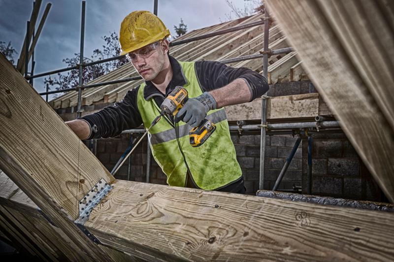 Person using a DEWALT cordless drill on wooden beams at a construction site, wearing safety gear and high-visibility vest.