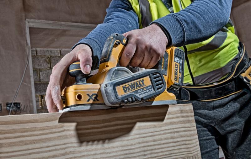 Person using a DEWALT DCP580N cordless planer on a wooden surface at a construction site.