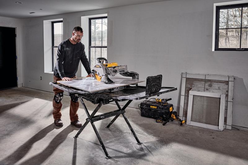 A person stands beside a DEWALT D36000S tile saw setup on its stand in a room under construction. The saw is placed on a foldable black stand, and there are tools and a DEWALT tool bag on the floor nearby. The room has unfinished walls, windows, and construction materials leaning against the wall.