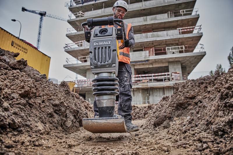 DEWALT compactor in use at a construction site, operated by a worker in safety gear, surrounded by soil and buildings.