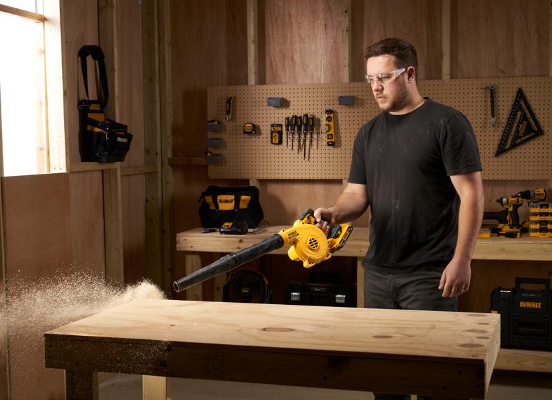 Person using a DEWALT DCV100 blower to clean sawdust off a wooden workbench in a workshop.