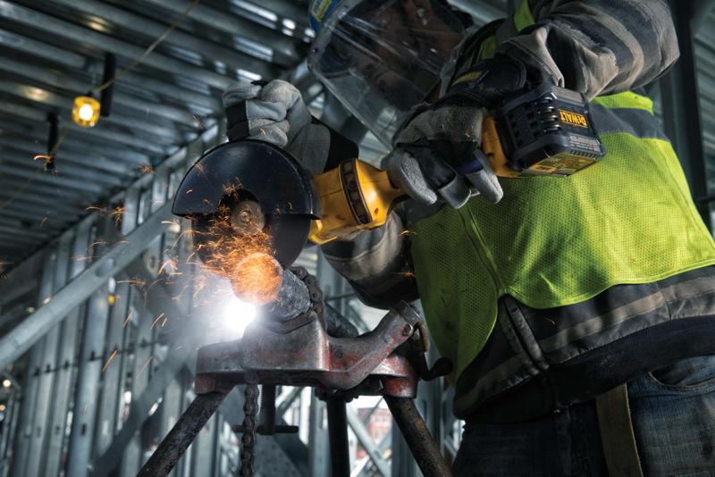 A close-up of a person wearing protective gloves and a safety vest using a DEWALT cordless angle grinder to cut metal, with sparks flying, inside an industrial construction site.