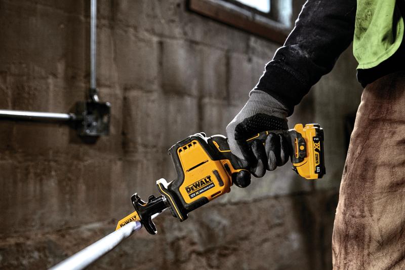 Gloved hand holding a DEWALT DCS312D2 cordless reciprocating saw cutting a metal pipe indoors.